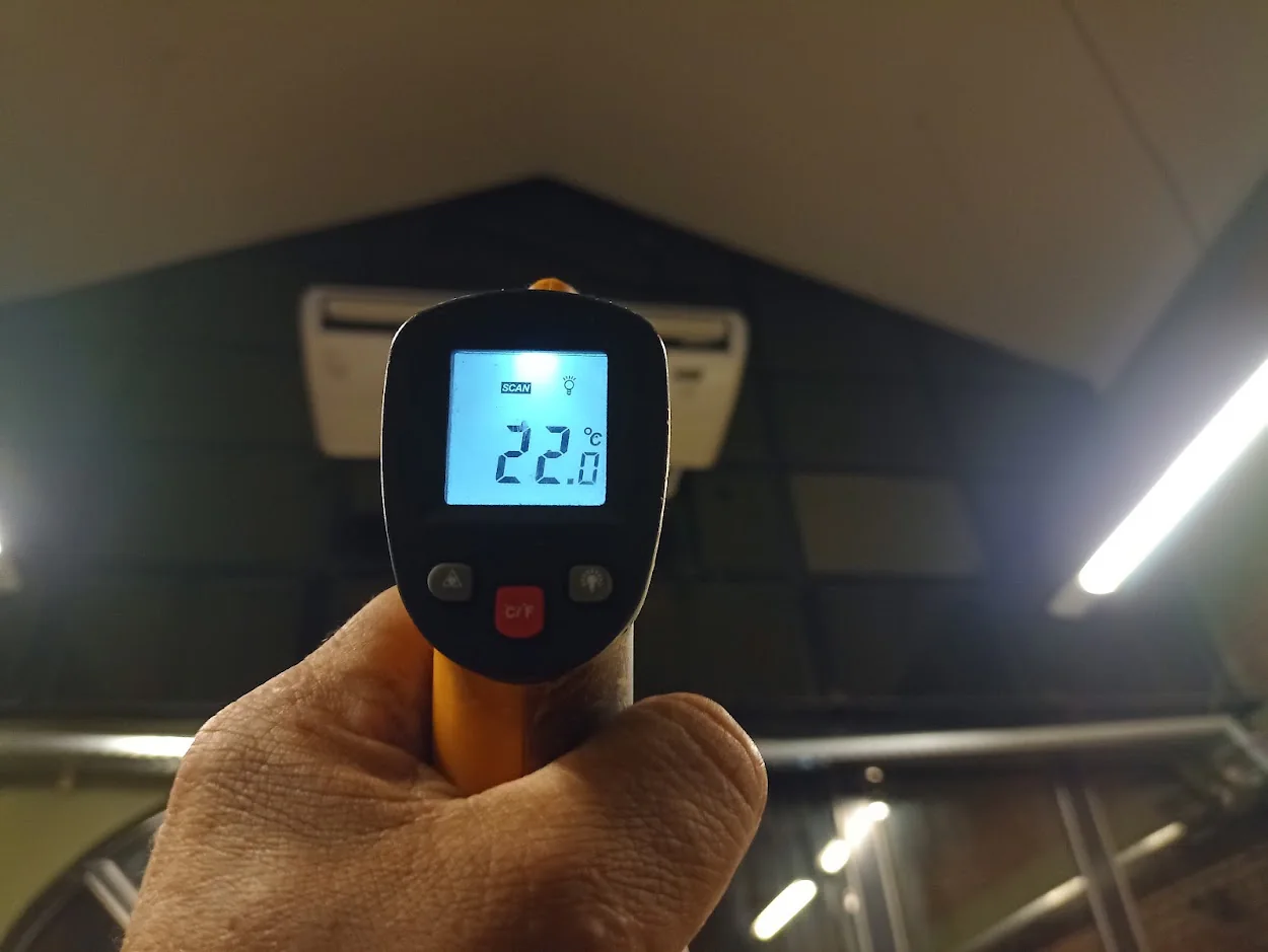 Mano sosteniendo un termómetro digital que muestra una lectura frente a un equipo de aire acondicionado.